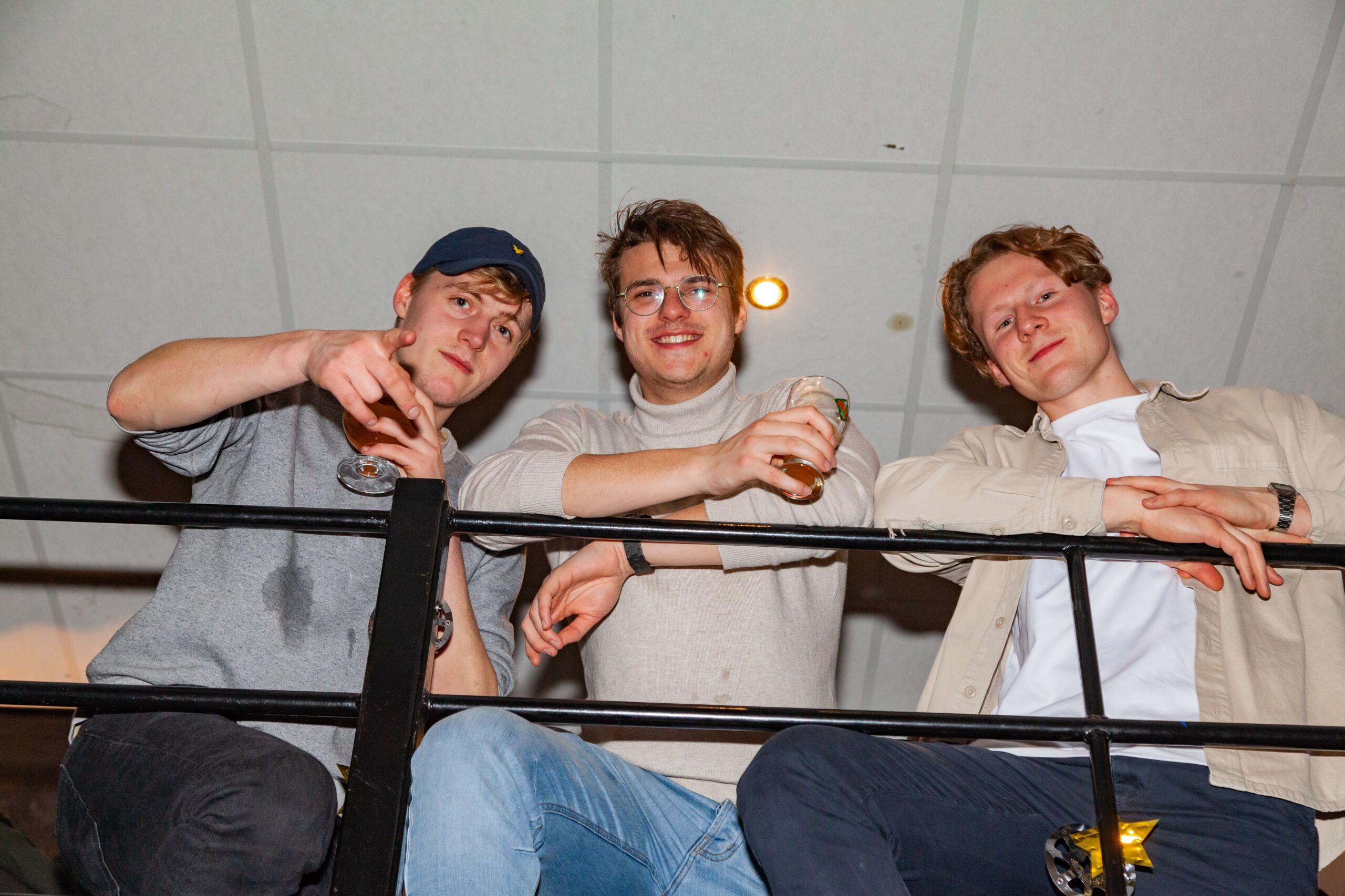 Drie jonge mannelijke studenten leunen op een reling en genieten van een gezellig samenzijn. De man links wijst naar de camera, terwijl de andere twee biertjes vasthouden en naar de kijker glimlachen. Ze zijn casual gekleed en lijken in een goede stemming te zijn, en dat op een normale donderdagavond op onze soos.