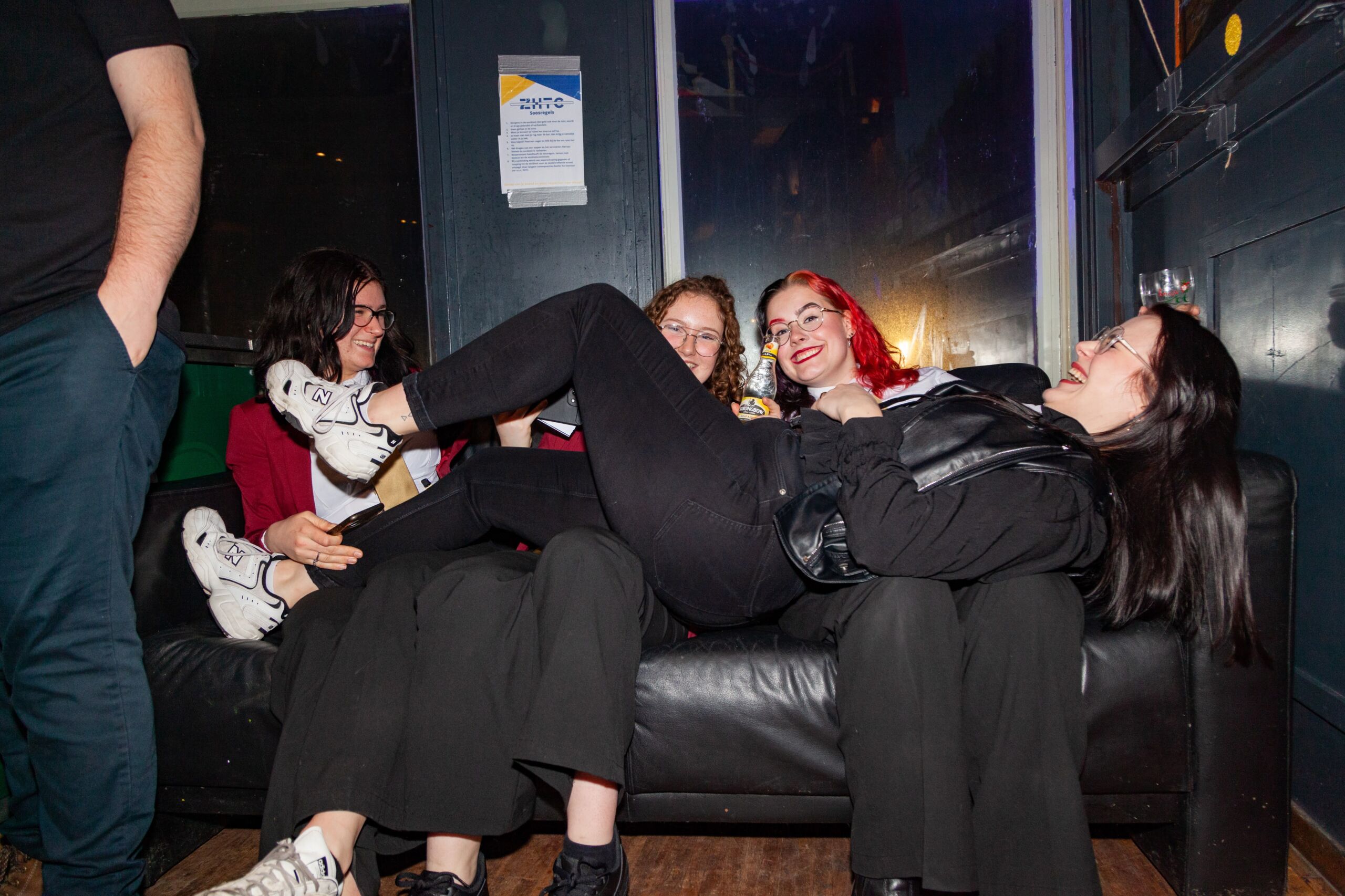 Een groep jonge vrouwen lacht en vermaakt zich op een zwarte leren bank in een ongedwongen binnensetting. Ze zijn gekleed in moderne, ontspannen kleding en gaan vrolijk met elkaar om, wat een zorgeloos moment onder vrienden belichaamt.