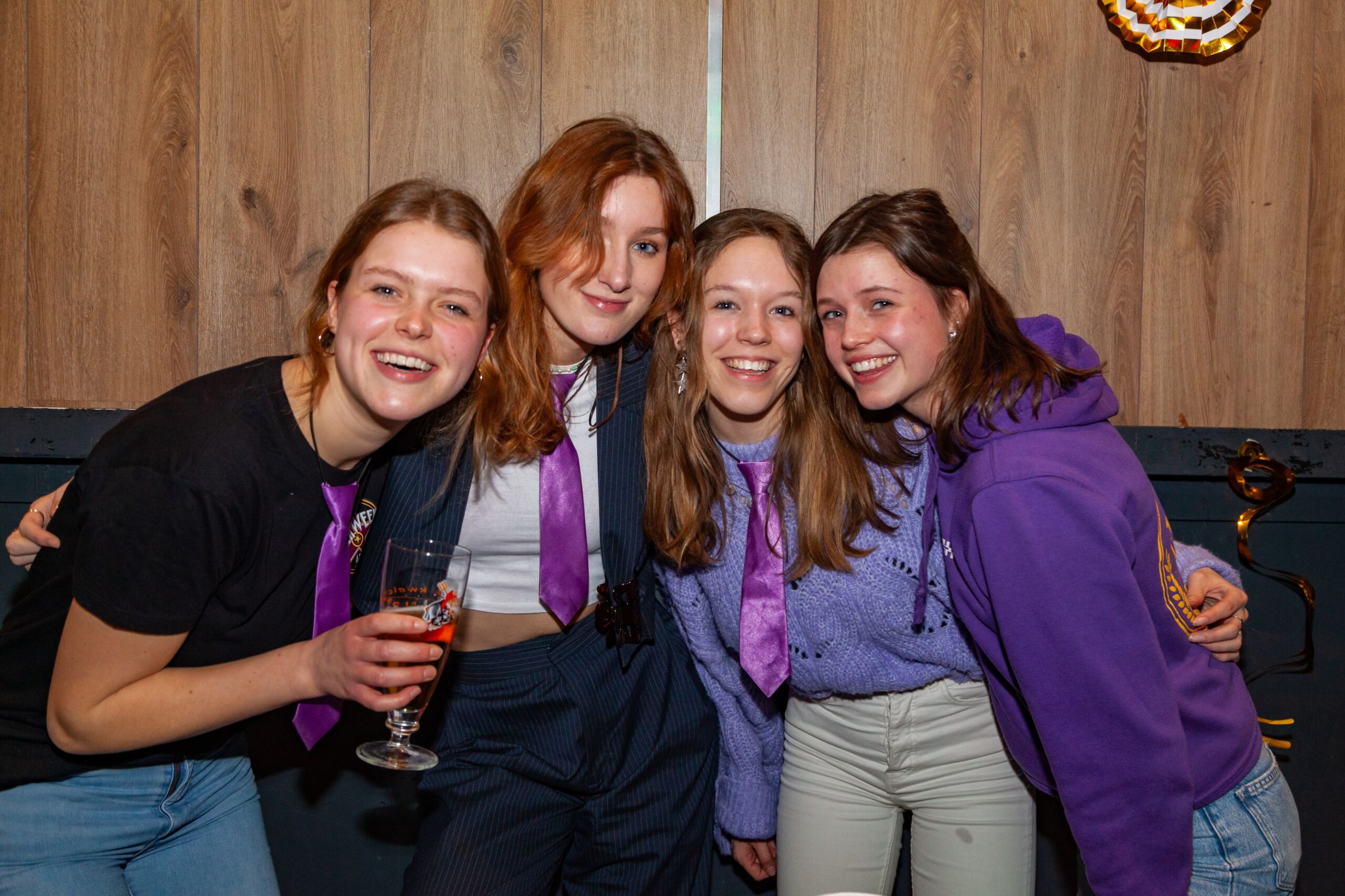 Vier vrouwlijke studenten vieren feest, mogelijk, dragen hun dispuutskleding. Ze houden drankjes vast en glimlachen hartelijk in een gezellige omgeving binnenshuis.