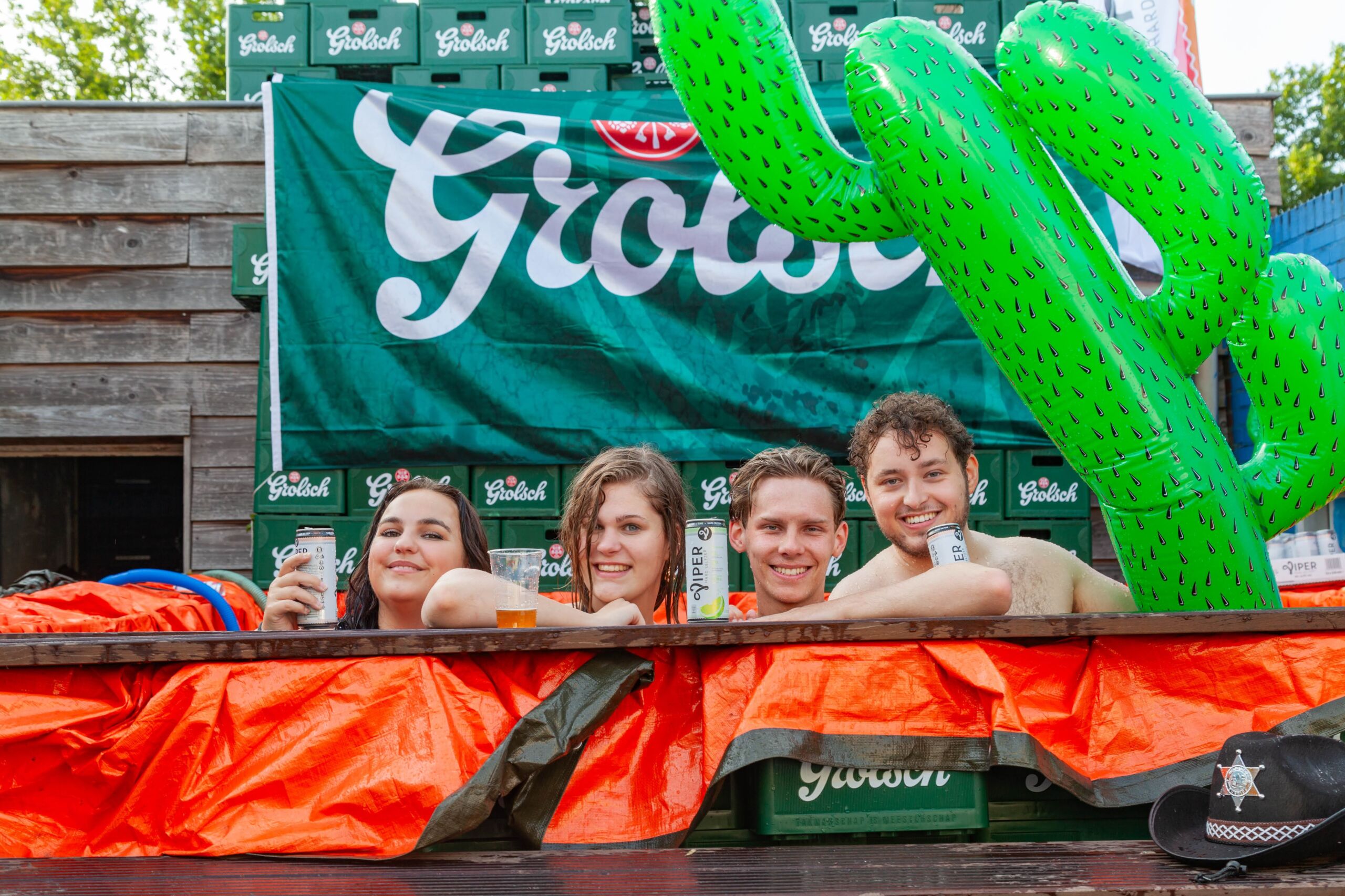 Vier vrienden in de tuin van onze soos, half ondergedompeld in een geïmproviseerd zwembad met Grolsch bier kistjes op de achtergrond. Ze lachen en houden drankjes vast, met een grote opblaasbare cactus die een speels tintje aan het tafereel geeft.