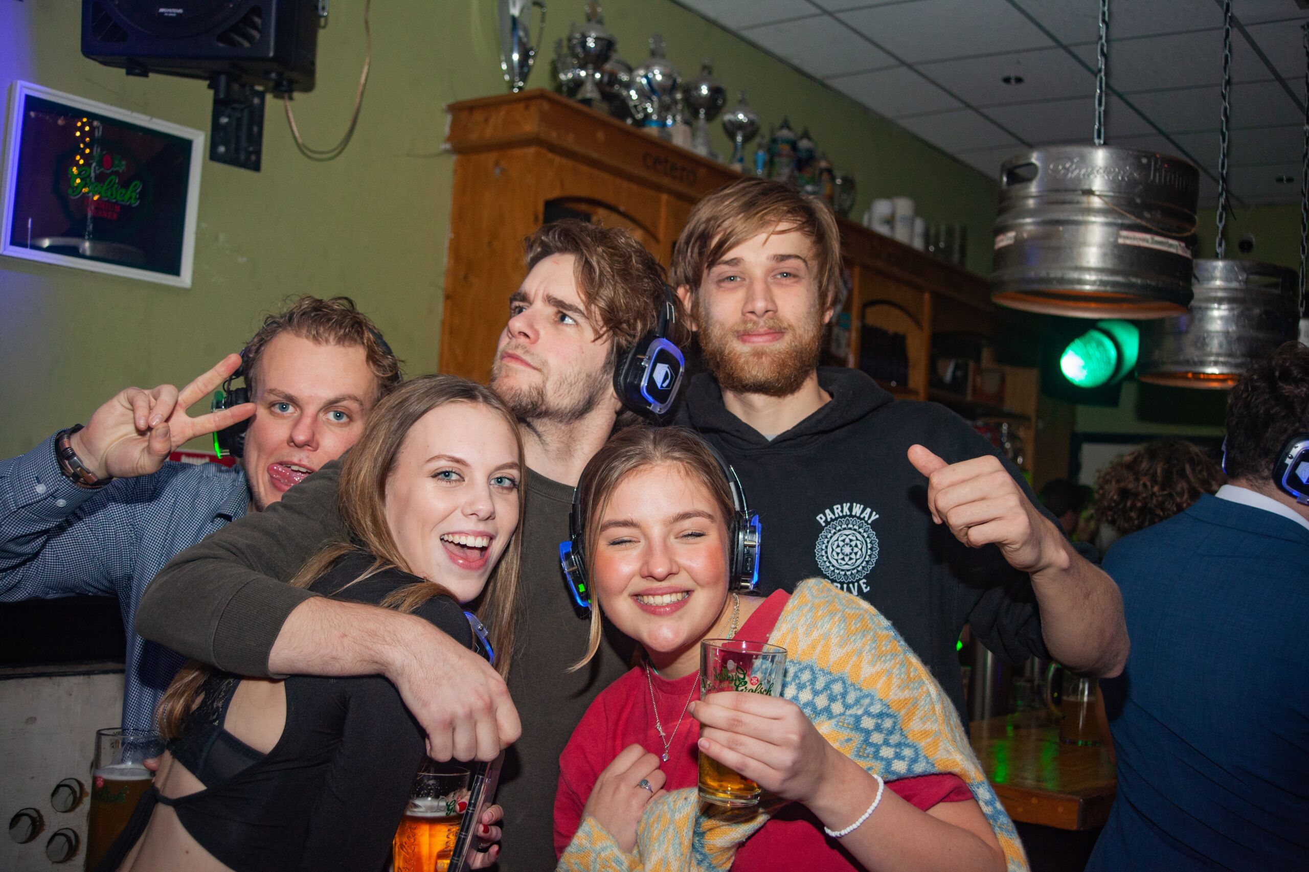 Een levendige groep van jongvolwassenen aan een bar, poserend met drankjes en speelse gebaren makend. Ze genieten van een levendige feestsfeer, met biervaten die aan het plafond hangen en feestelijke verlichting op de achtergrond.