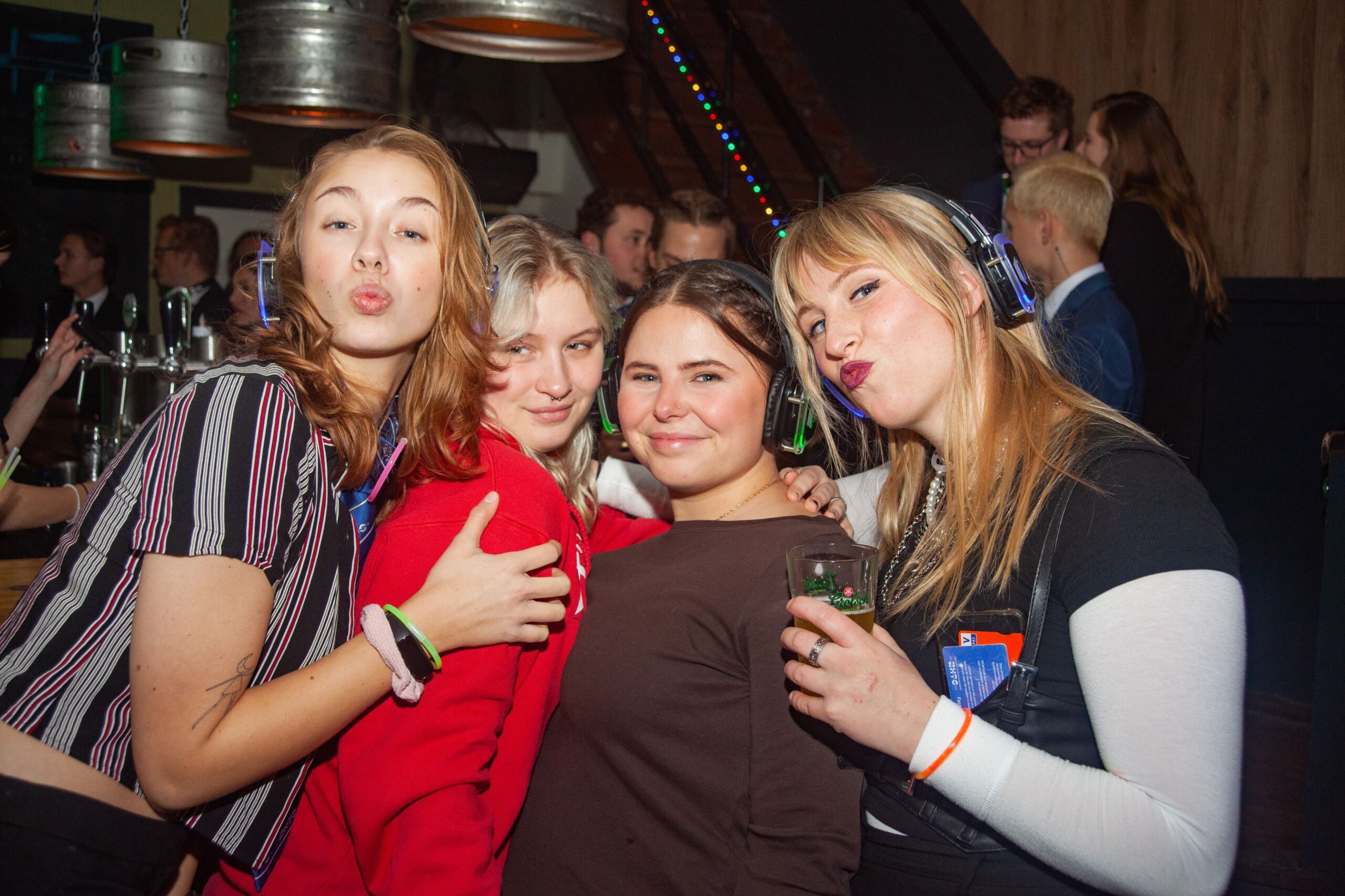Vier vrouwlijke studenten knuffelen en poseren voor een foto op een feestje. Een van hen tuit speels haar lippen, terwijl de anderen glimlachen en drankjes vasthouden. De setting bestaat uit feestverlichting en biervaten op de achtergrond, wat een levendige sfeer creëert.