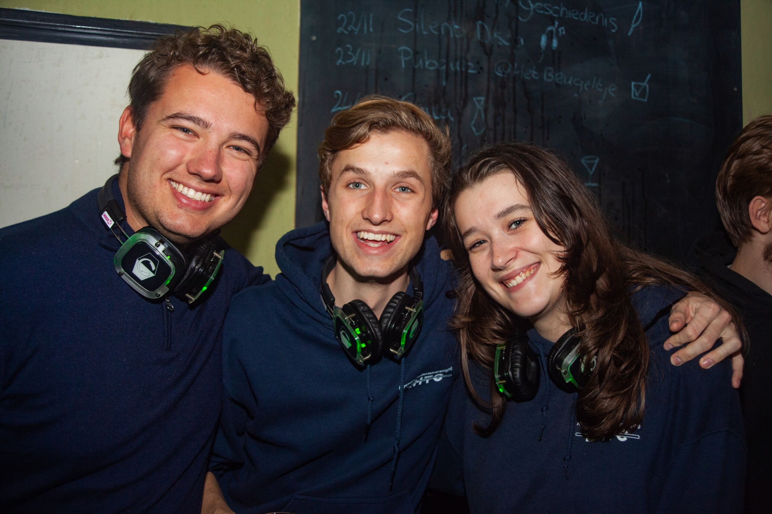 Drie studenten die onze ZHTC- weatshirts dragen en over-ear koptelefoons om hun nek. Ze glimlachen en poseren samen, wat een levendige sociale sfeer weerspiegelt.