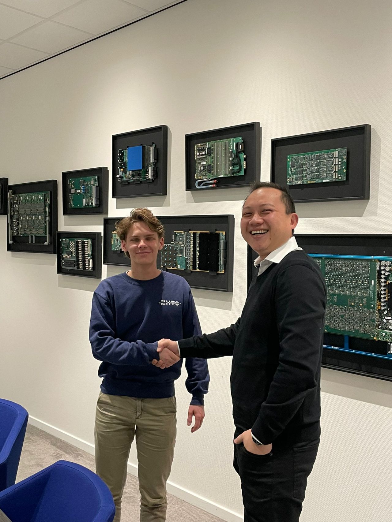 Two men smiling and shaking hands in an office, with one wearing a casual sweatshirt and the other in a black jacket, against a wall decorated with various computer circuit boards framed as art.